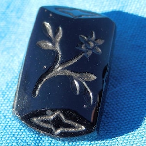 Puede incluir: Un camafeo de vidrio negro con un diseño tallado de una flor y un tallo. El camafeo es rectangular y tiene una superficie lisa y pulida.