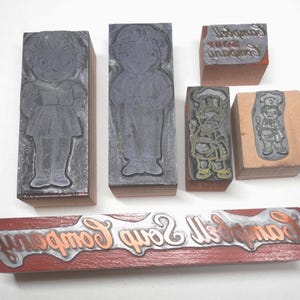 Puede incluir: Una colección de bloques de impresión vintage. Los bloques presentan imágenes de un niño, un chef y el texto "Campbell's All Soup Company". Los bloques están hechos de madera y metal, con las imágenes y el texto en relieve.