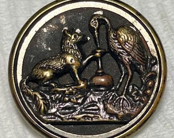 May include: A vintage button with a gold rim and a black background. The button features a detailed image of a fox and a crane standing on a pedestal.