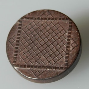 Könnte beinhalten: Ein brauner, runder Knopf mit geometrischem Muster. Der Knopf hat ein quadratisches Muster mit einem Gitter aus kleinen Quadraten im Inneren.