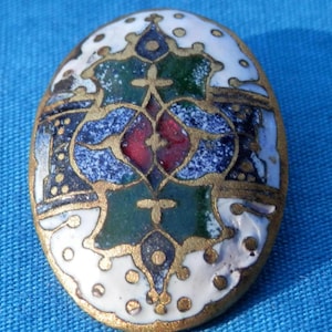 May include: Oval-shaped button with a white background and a colorful geometric design. The design features blue, green, red, and gold accents.