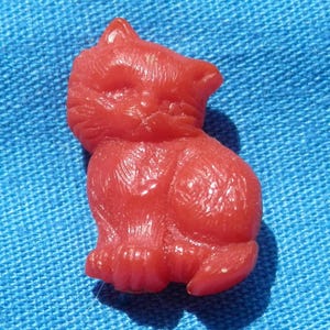 Puede incluir: Una pequeña figura de gato rojo en posición sentada. El gato tiene una superficie texturizada detallada y está sobre un fondo azul. La figura tiene una oreja puntiaguda y una pequeña cola.