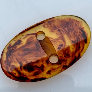 Puede incluir: Un botón ovalado de color marrón y amarillo con dos agujeros. El botón tiene un patrón de mármol.