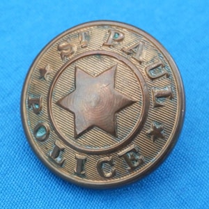 Könnte beinhalten: Ein Vintage-Polizeiknopf aus Bronze aus St. Paul, Minnesota. Der kreisförmige Knopf zeigt die Worte "ST. PAUL POLICE" um einen sechsspitzigen Stern. Der Knopf hat ein leicht gealtertes Aussehen.