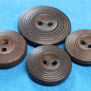 Könnte beinhalten: Vier braune, kreisförmige Knöpfe mit je zwei Löchern, angeordnet auf einem blauen Stoff. Die Knöpfe haben ein geriffeltes, konzentrisches Design. Die Knöpfe variieren leicht in der Größe, der größte Knopf befindet sich oben.