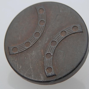 Könnte beinhalten: Ein dunkelgrauer Metallknopf mit einem dekorativen Design aus zwei geschwungenen Linien mit Kreisen an den Rändern.