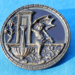 May include: A round, antique-style button with a detailed relief design. The button features a cherubic figure with wings, a fountain, and decorative elements. The button has a bronze finish and a textured edge.