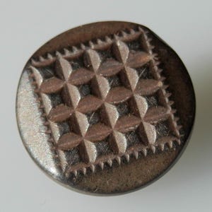 Könnte beinhalten: Ein runder, brauner Metallknopf mit geometrischem Muster. Der Knopf hat ein erhabenes, strukturiertes Muster aus Quadraten und Rauten.