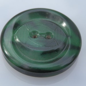 Puede incluir: Un botón de plástico verde con dos agujeros. El botón tiene un patrón de espiral y un centro elevado y texturizado.