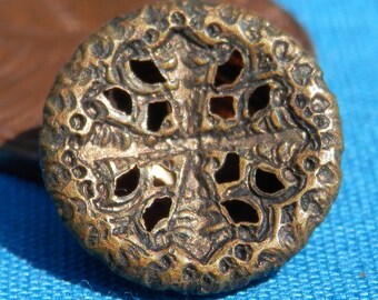 May include: Close-up of a round, antique brass button with an ornate, openwork design. The button features a central cross-like pattern with decorative details and small holes. The button is on a blue surface.
