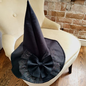 May include: A black witch's hat with a large black bow and sparkly trim. The hat is sitting on a chair.