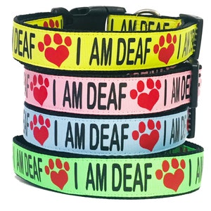 May include: Four dog collars stacked on top of each other. The collars are made of black webbing with a red paw print and heart design. The text "I AM DEAF" is printed in black on each collar. The collars are available in yellow, pink, blue, and green.