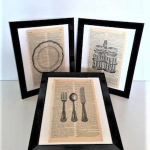 May include: Three framed art prints featuring black ink illustrations on vintage dictionary pages. One print shows a dinner plate, another displays salt and pepper shakers, and the third depicts a fork, spoon, and knife set in black frames.