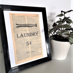 May include: A framed vintage-style print with the word "LAUNDRY" in large letters. The print is set against a cream-colored background and framed in a black frame. A small houseplant in a white pot sits to the right.