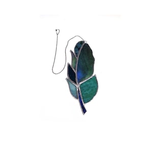 May include: A stained glass feather ornament featuring various shades of blue, teal, and green glass pieces joined by silver-toned metal solder lines. The feather is suspended from a thin silver-colored metal chain attached to the top edge.