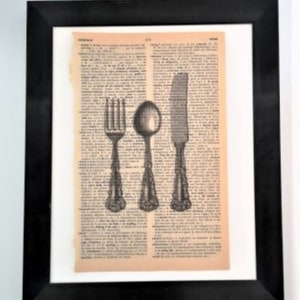 May include: A framed wall art piece featuring a vintage dictionary page with a black ink illustration of a fork, a spoon, and a knife centered on the paper. The artwork is enclosed in a simple black frame with a white mat border surrounding the page.