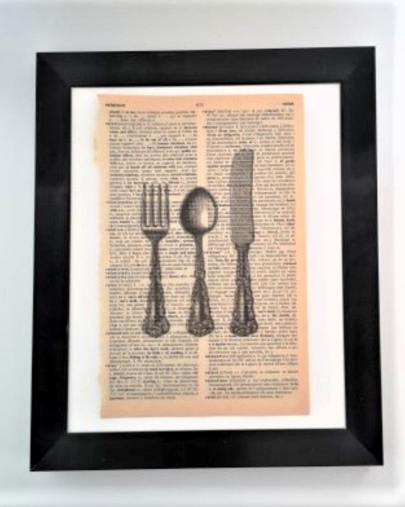 May include: A framed wall art piece featuring a vintage dictionary page with a black ink illustration of a fork, a spoon, and a knife centered on the paper. The artwork is enclosed in a simple black frame with a white mat border surrounding the page.