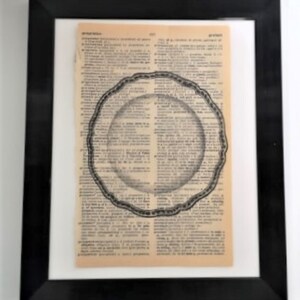 May include: A framed wall art piece featuring a vintage dictionary page with aged, tan paper. Centered on the text is a black and white line drawing of a round, scalloped-edge plate. The entire artwork is enclosed in a simple black frame.
