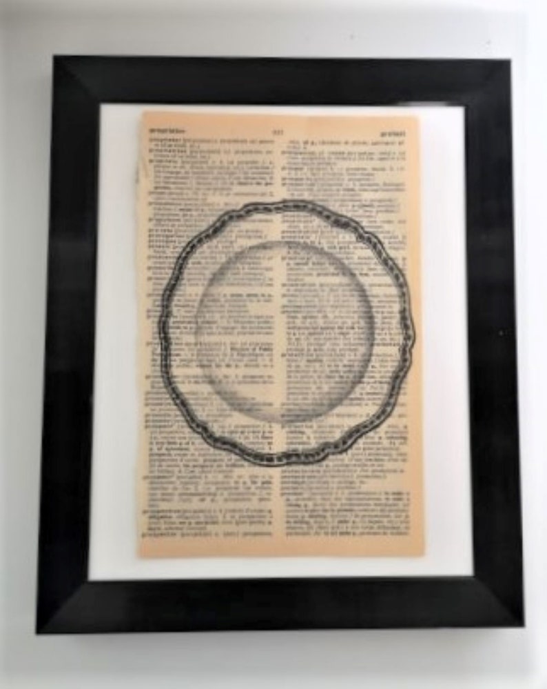 May include: A framed wall art piece featuring a vintage dictionary page with aged, tan paper. Centered on the text is a black and white line drawing of a round, scalloped-edge plate. The entire artwork is enclosed in a simple black frame.