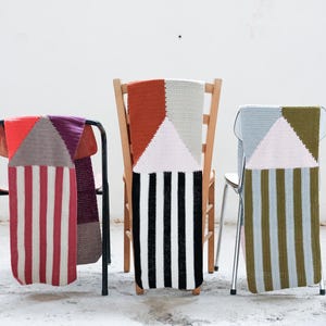 Op de afbeelding: Drie stoelen met patronen stoelhoezen. De hoezen tonen geometrische ontwerpen met driehoeken en strepen in rood, oranje, paars, zwart, wit en groen. De stoelen staan tegen een witte muur.