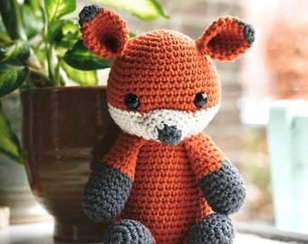 May include: A crocheted orange fox plush toy with gray accents on the ears, paws, and nose. The toy is sitting on a white surface.