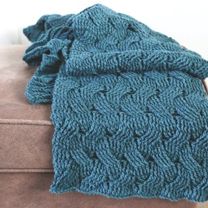 Op de afbeelding: Een tealblauwe gebreide deken met een kabelbreipatroon. De deken is gevouwen en gedrapeerd over een bruine ottomane.