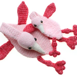 May include: Two crocheted pink flamingo toys. The larger flamingo has a smaller flamingo on its back. Both have red wings and legs.