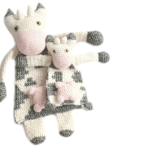 May include: A crocheted stuffed animal toy in the shape of a cow, with a smaller crocheted stuffed animal toy in the shape of a cow inside the larger cow's arms. The larger cow is white with black and gray spots, and the smaller cow is white with black spots. Both cows have pink noses and eyes.