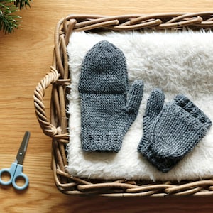 Op de afbeelding: Een paar grijze gebreide vingerloze handschoenen in een rieten mand met een witte nep bontvoering.