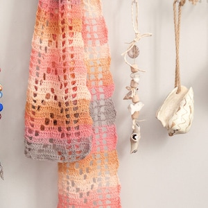 May include: A collection of handmade items hanging on a white wall. A crocheted scarf in shades of pink, orange, and gray hangs on the left. A beaded necklace with silver charms hangs next to it. On the right, a white seashell hangs from a string, and a brown bar of soap hangs from another string.
