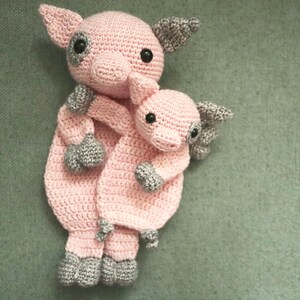 May include: Two crocheted pink pig plushies, one larger than the other, are hugging. The smaller pig is nestled in the arms of the larger pig. Both pigs have gray crocheted noses and feet.