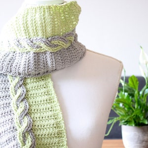 Op de afbeelding: Een gehaakte sjaal met een gevlochten ontwerp in grijs en groen. De sjaal wordt getoond op een wit mannequin.