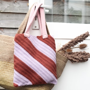 Könnte beinhalten: Eine gehäkelte Tragetasche mit einem diagonalen Streifenmuster in Lila- und Brauntönen. Die Tasche hat rosa Henkel und hängt an einer Tür.