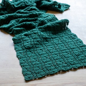 Pode incluir: Uma echarpe de crochê verde com um padrão de renda. A echarpe está dobrada e colocada sobre uma superfície de madeira.
