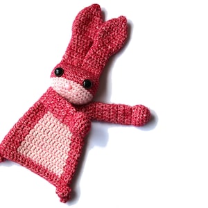 May include: A pink and red crocheted bunny toy with black eyes. The bunny is lying on its side with its arm outstretched.