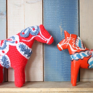 May include: Two Dala horses, one crocheted and one wooden. The crocheted horse is red with blue and white patterns. The wooden horse is red with white and blue floral designs. They are displayed against a wooden backdrop.