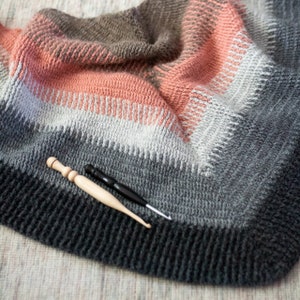 Könnte beinhalten: Nahaufnahme einer gehäkelten Decke mit einem Farbverlauf von Grau, Weiß, Rosa und Braun. Zwei Häkelnadeln, eine aus Holz und eine schwarze, liegen auf der Decke.