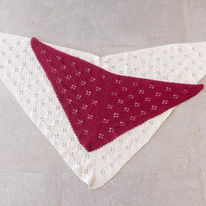 Könnte beinhalten: Zwei dreieckige Stricktücher mit einem sich wiederholenden Lochmuster. Ein Tuch ist cremeweiß und das andere ist in einem tiefen Magentarot gehalten. Beide Artikel haben eine spitz zulaufende Unterkante und einen breiten, fein gewellten Rand.