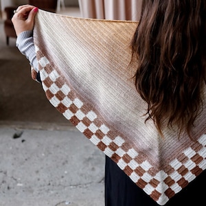 May include: A knitted shawl in a gradient of beige to brown hues. The shawl has a checkered border in brown and white. The design is a blend of textures and colours, creating a visually appealing accessory.
