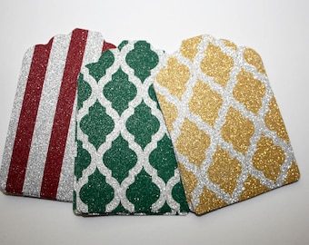 May include: Three gift tags with glitter. One tag has red and white stripes, one has a green and white geometric pattern, and one has a gold and white geometric pattern.