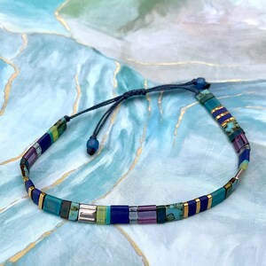 May include: A beaded bracelet with a sliding knot closure. The bracelet features rectangular beads in shades of blue, green, purple, and gold. The beads are strung on a dark blue cord, creating a colorful and stylish accessory.