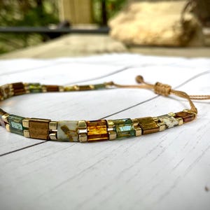 May include: A beaded bracelet with a sliding knot closure. The bracelet features a pattern of small, rectangular beads in shades of brown, green, gold, and white. The beads are strung together with a tan cord.