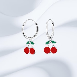 May include: A pair of silver hoop earrings with red cherry charms. Each charm features two red cherries with green leaves. The earrings are displayed against a soft, white background.