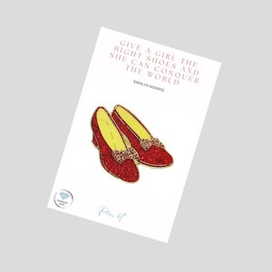 May include: Red glitter ruby slipper enamel pin with a gold bow and yellow lining. The pin features the quote "Give a girl the right shoes and she can conquer the world" by Marilyn Monroe.