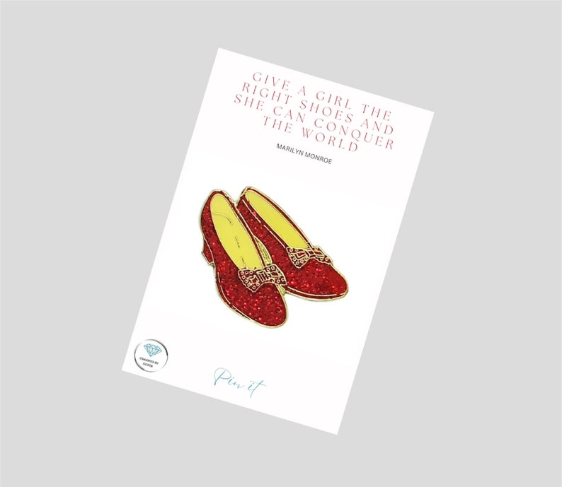 May include: Red glitter ruby slipper enamel pin with a gold bow and yellow lining. The pin features the quote "Give a girl the right shoes and she can conquer the world" by Marilyn Monroe.