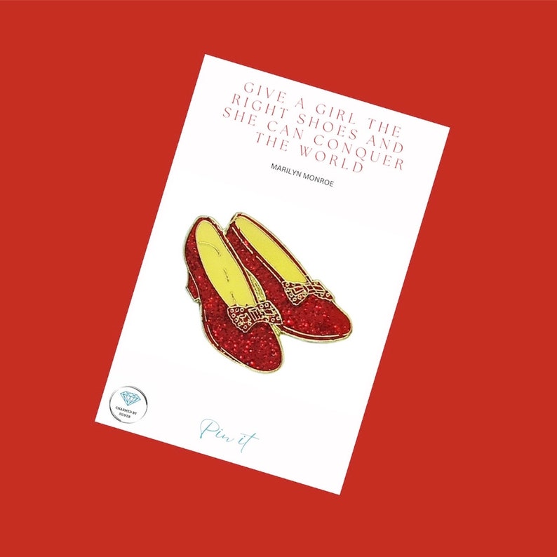 May include: Red glitter enamel pin of a pair of ruby slippers with a gold buckle and bow. The pin features the quote "Give a girl the right shoes and she can conquer the world" by Marilyn Monroe.