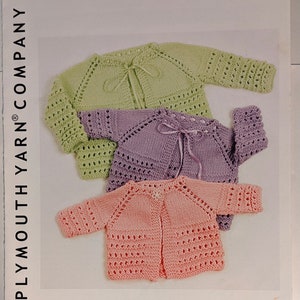 May include: Three knitted baby jackets in green, purple, and pink. The jackets are made with a lace stitch and have a tie closure at the neck. The image includes the text "Encore® Worsted 1354" and "PLYMOUTH YARN® COMPANY" and "TOP DOWN BABY JACKET".