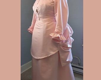 May include: A light pink two-piece ensemble consisting of a long-sleeved bodice with a high collar and a row of small buttons down the front, paired with a matching floor-length skirt featuring a draped bustle effect and ruffled sleeve cuffs.