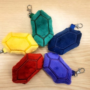 May include: Six plush keychains shaped like gemstones in different colors: yellow, green, blue, red, purple, and blue. Each keychain has a silver metal ring.