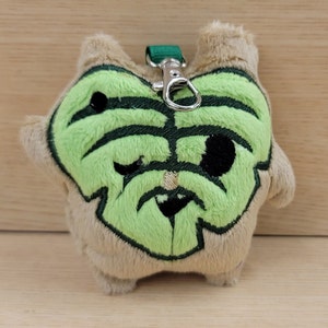 May include: A plush keychain shaped like a green leaf with a brown furry body. The leaf has a black eye and a white nose.